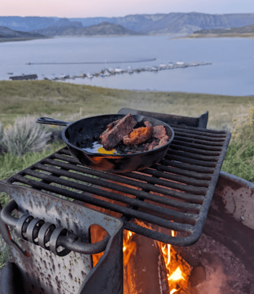 A cast iron skillet with cooking meat sits on a grill by a lake, surrounded by hills at sunset.
