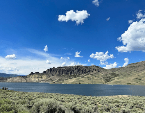A serene lake surrounded by green hills and rocky cliffs under a bright blue sky with fluffy white clouds.