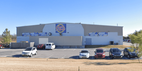 Exterior of a roller skating rink with a large sign, surrounded by a parking lot filled with cars. Clear blue sky.