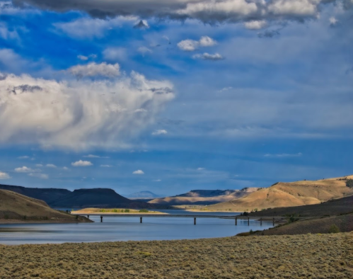 A serene landscape featuring a river, rolling hills, and a bridge under a blue sky with scattered clouds.