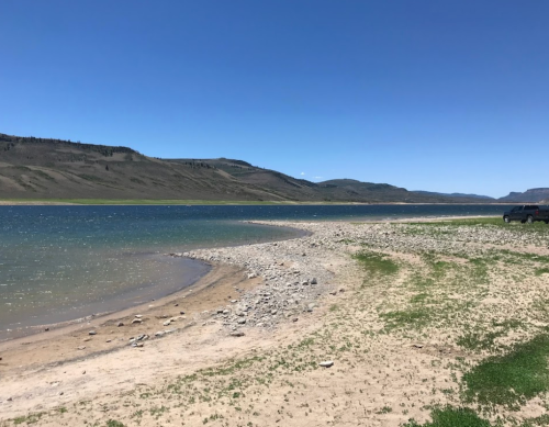 A serene lakeside view with sandy shores, rocky edges, and distant hills under a clear blue sky.