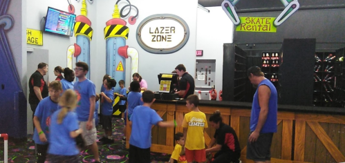 A busy indoor entertainment center with kids in blue shirts, a skate rental area, and staff assisting at the counter.