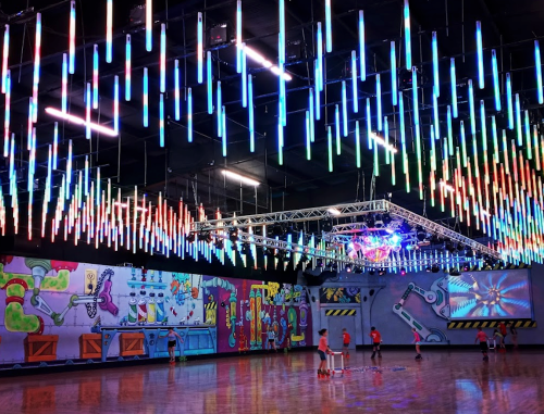 A vibrant indoor skating rink with colorful LED lights hanging from the ceiling and graffiti art on the walls.