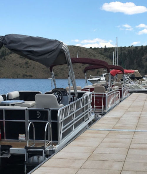 A row of parked pontoon boats at a marina, with a scenic lake and hills in the background.