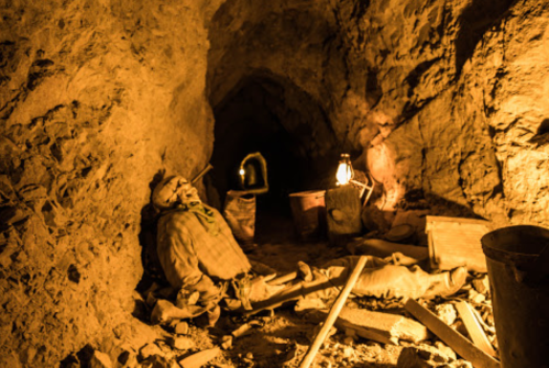 A dimly lit mine tunnel with a skeleton resting on the ground, surrounded by mining tools and lanterns.
