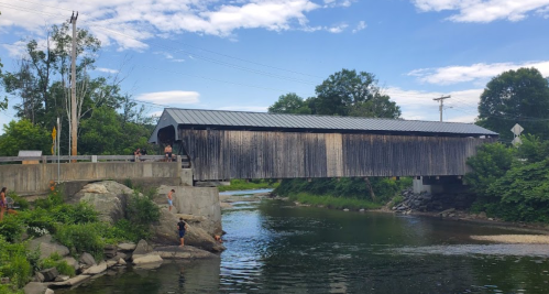 A wooden covered bridge spans a river, surrounded by greenery and people enjoying the water on a sunny day.