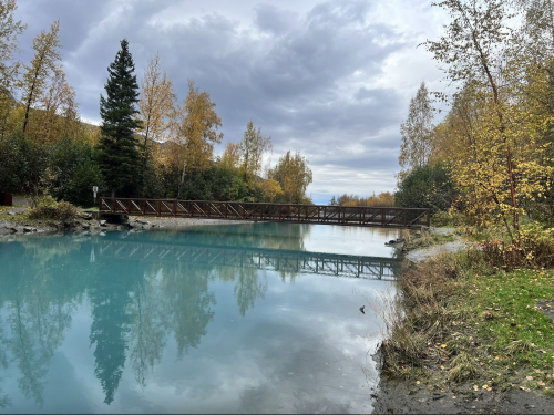 A tranquil river scene with a wooden bridge, surrounded by autumn foliage and cloudy skies. Reflections in the water.