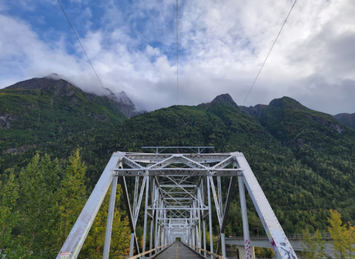 A white metal bridge leads towards lush green mountains under a cloudy sky.