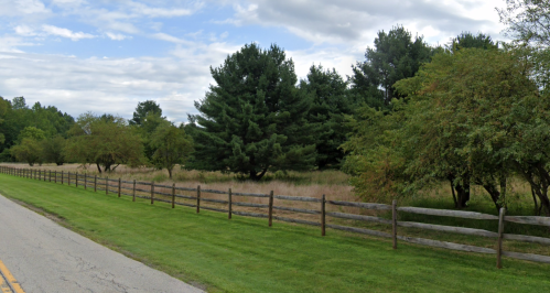 A grassy area with a wooden fence, trees, and a cloudy sky in the background along a roadside.