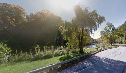 A scenic view of a river bordered by trees, with a winding road alongside and sunlight shining through the foliage.