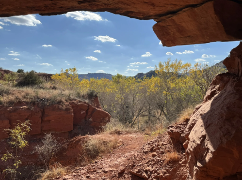 View through a rocky opening, showcasing a landscape of trees with yellow leaves under a blue sky with scattered clouds.