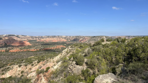 A panoramic view of rugged red rock formations and green vegetation under a clear blue sky.