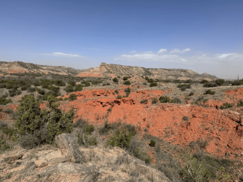 A scenic view of red rock formations and green shrubs under a clear blue sky in a rugged landscape.