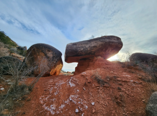 A large boulder balanced on a smaller rock, set against a cloudy sky and reddish soil. Sparse vegetation surrounds the scene.