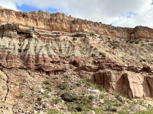 Layered rock formations with reddish and gray hues under a partly cloudy sky. Vegetation scattered at the base.
