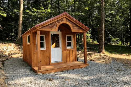 A small wooden cabin with a porch, surrounded by trees and gravel, set in a natural outdoor environment.