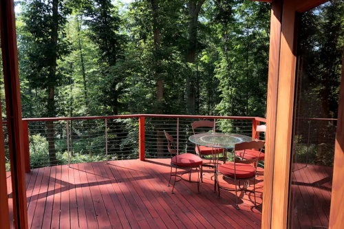 A wooden deck with a glass table and chairs, surrounded by lush green trees in a serene forest setting.