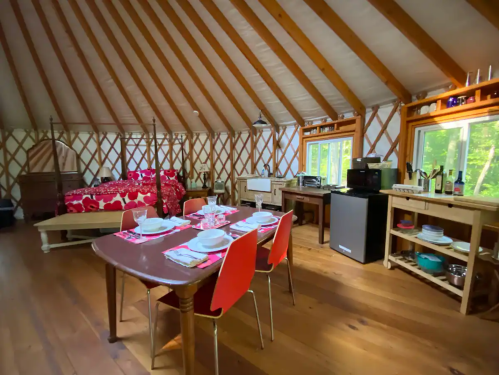 Cozy yurt interior with a dining table, kitchenette, and a bed, featuring wooden beams and natural light.