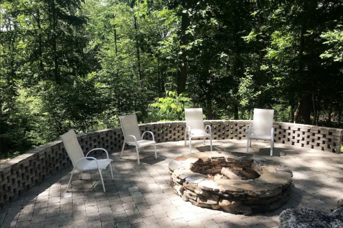 A stone fire pit surrounded by white chairs in a wooded outdoor setting.