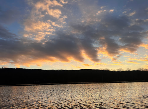 A serene sunset over a calm lake, with colorful clouds reflecting on the water and a dark silhouette of hills in the background.