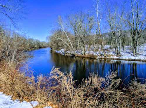 A serene river flows through a winter landscape, surrounded by bare trees and patches of snow under a clear blue sky.