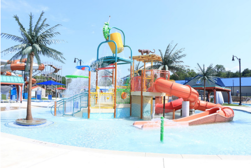 Colorful water play structure with slides and fountains in a sunny pool area, surrounded by palm trees.