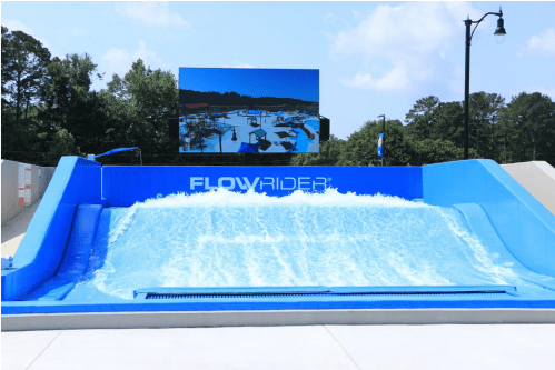 A FlowRider surf simulator with a large screen in the background, surrounded by trees and blue skies.