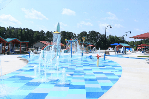 A colorful splash pad with water features and blue checkered flooring, surrounded by trees and sunny skies.