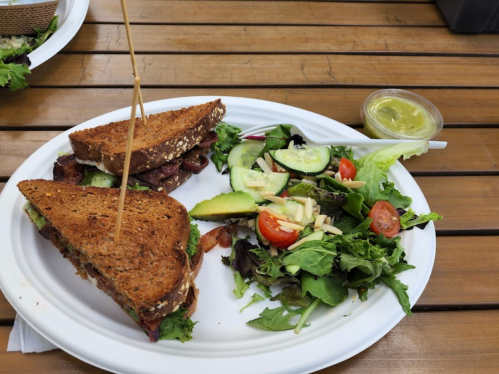 A plate with a sandwich on whole grain bread and a side salad with tomatoes, cucumbers, and a small container of dressing.