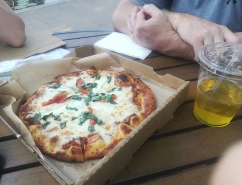 A pizza with cheese, tomatoes, and basil sits in a box on a table next to a drink and hands.