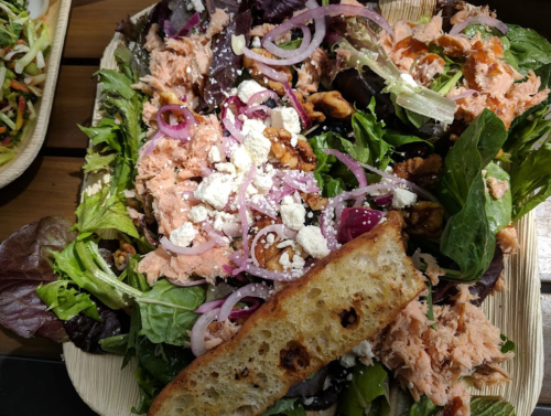 A vibrant salad with mixed greens, shredded salmon, red onions, walnuts, feta cheese, and a slice of toasted bread.