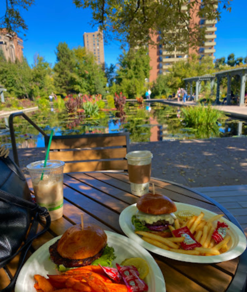 A table with two plates of burgers and fries, drinks, overlooking a serene pond and greenery in a park.