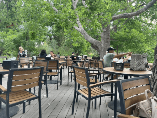 Outdoor café scene with wooden tables and chairs, surrounded by greenery and people enjoying their time.