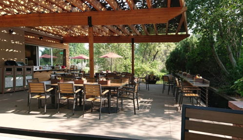Outdoor dining area with wooden tables and chairs, shaded by a pergola, surrounded by greenery and umbrellas.