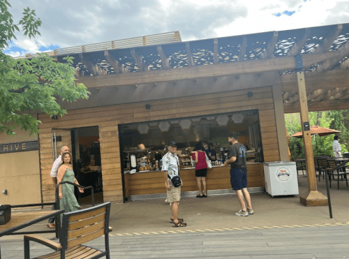 A wooden café with outdoor seating; people are ordering and enjoying drinks on a sunny day.