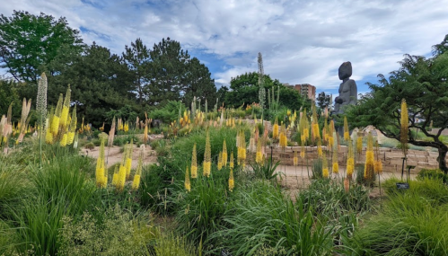 A serene garden with tall yellow flowers, lush greenery, and a large statue in the background under a cloudy sky.