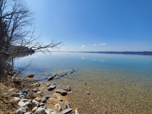 A serene lake scene with clear water, rocky shore, and trees under a bright blue sky.