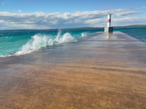 A pier extends over turquoise waters, with waves crashing against it and a red and white lighthouse in the background.
