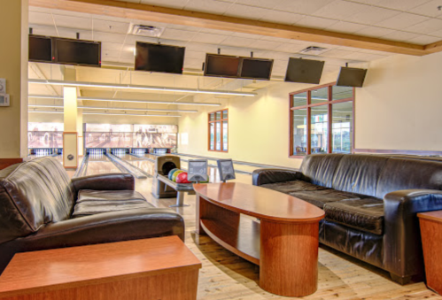A cozy bowling alley lounge with leather sofas, a wooden table, and bowling lanes visible in the background.