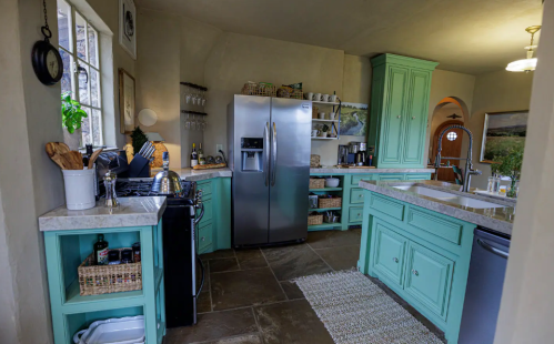 A cozy kitchen with mint green cabinets, stainless steel appliances, and a stone floor, featuring natural light and decor.