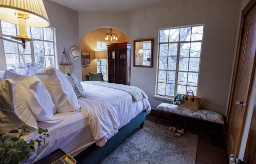Cozy bedroom with a large bed, natural light from windows, and a view of trees outside. Warm, inviting decor.