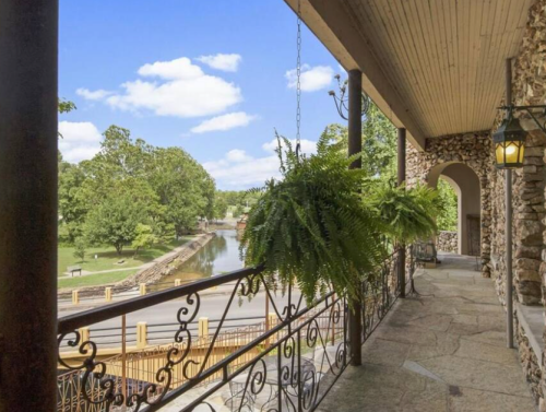 A scenic view from a porch with hanging ferns, overlooking a river and lush greenery under a blue sky.