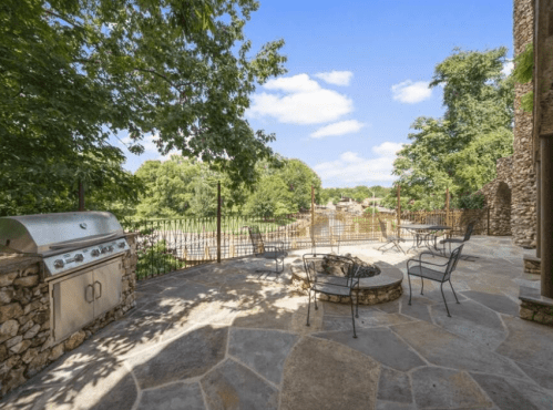 Outdoor patio with a grill, fire pit, and seating, surrounded by trees and a scenic view. Bright blue sky above.