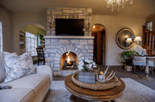 Cozy living room with a stone fireplace, TV above, elegant decor, and a round coffee table with flowers.