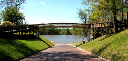 A wooden bridge spans over a calm river, surrounded by green trees and a clear blue sky.