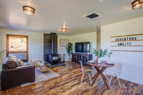 Cozy living area with a sofa, TV, dining table, and a wood stove, featuring a sign that says "Adventure Awaits."