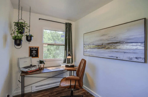 A cozy workspace featuring a desk, chair, plants, and a record player, with a large abstract painting on the wall.