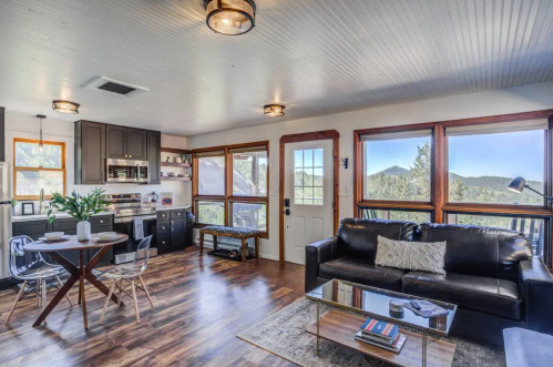 Cozy interior with a black sofa, wooden dining table, and large windows showcasing mountain views.