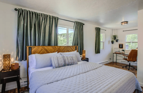 Cozy bedroom with a large bed, decorative pillows, green curtains, and a workspace with a desk and chair by the window.