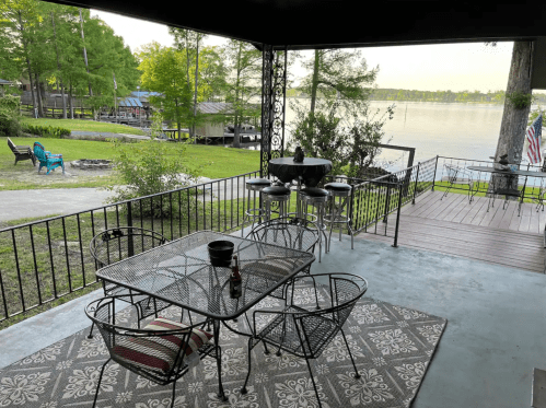 A lakeside patio with a table, chairs, and a view of the water and greenery in the background.